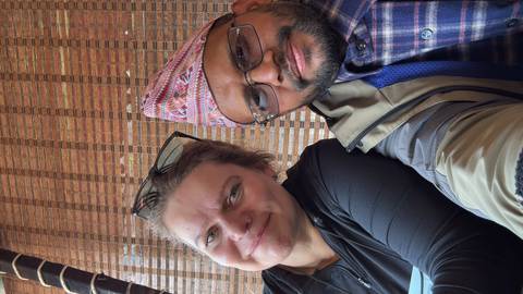 Close-up selfie of two smiling travellers, one wearing a traditional Nepali topi hat.