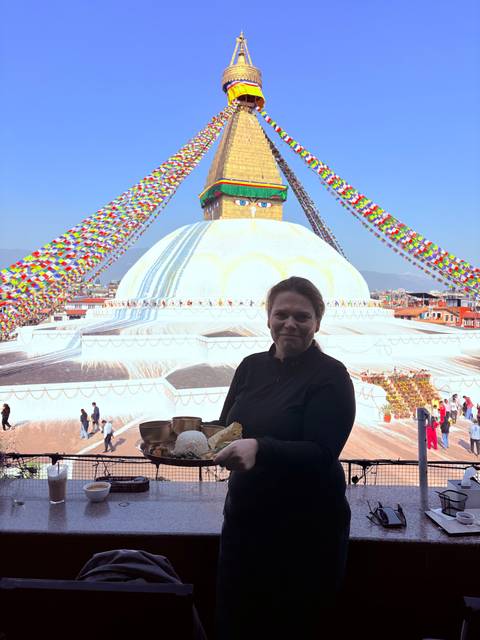 Woman holding a tray of Nepali food in front of the colorful Boudhanath Stupa draped with prayer flags.
