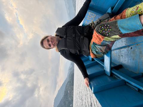 Woman sits relaxed in a blue wooden boat on a calm lake with misty Himalayan hills beyond.
