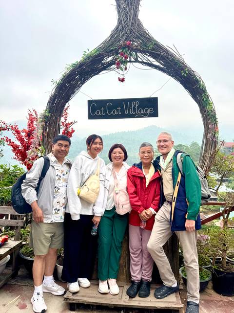 Travellers smile beneath the wooden Cat Cat Village arch amid misty mountain scenery.