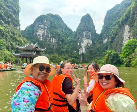 Smiling tourists in bright life vests ride a small boat through green karst cliffs and a riverside pagoda.