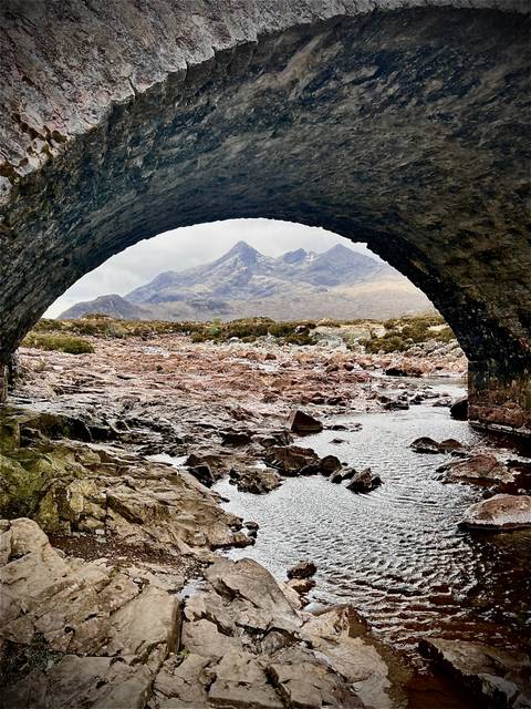 Rugged mountain peaks of the Cuillin framed beneath an old stone bridge arch with a rocky riverbed in front.