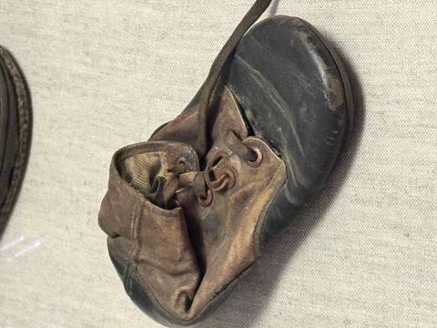 A worn, child-sized leather shoe displayed on a neutral fabric surface in a museum exhibit.