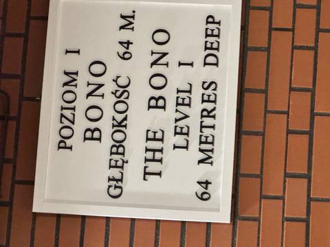 Bilingual depth-level sign in Polish and English mounted on a brick wall inside a tourist site.