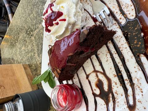 Plated slice of rich chocolate cake topped with berry sauce and whipped cream on a café table.