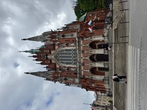 Tall red-brick gothic church with spired towers rising against a cloudy sky, pedestrians at the steps.