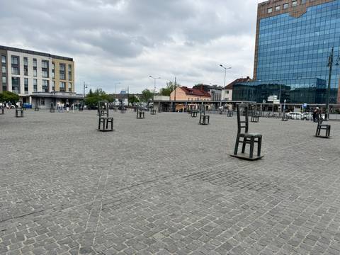Large cobbled square scattered with oversized metal chair sculptures, modern buildings around.