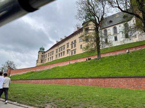 Long castle facade on a grassy hillside with brick retaining wall on a cloudy day.