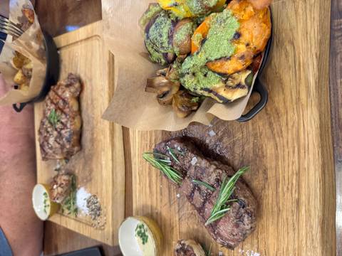 Grilled steak garnished with rosemary alongside roasted vegetables coated in green sauce on wooden boards.