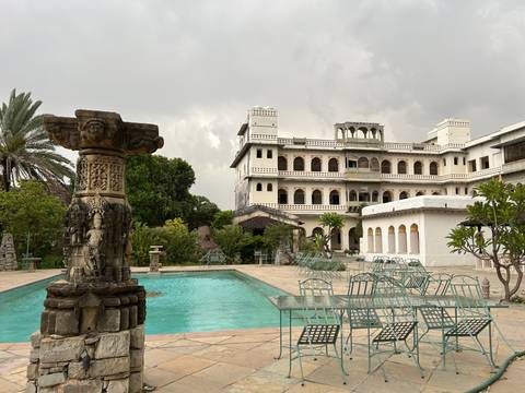 Heritage palace hotel with pool and metal garden furniture under an overcast monsoon sky.
