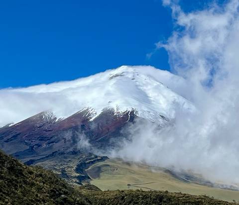 Snow-capped Cotopaxi volcano peak emerges dramatically from clouds against a deep blue sky.