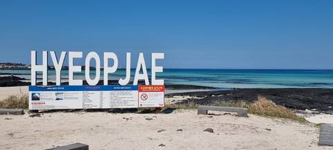 White Hyeopjae Beach sign on white sand with turquoise sea and blue sky beyond