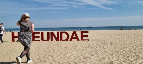 Tourist walking past red Haeundae beach sign on wide sandy shore with calm sea