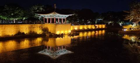 Nighttime reflection of traditional pavilion and stone wall in tranquil pond illuminated by warm lights