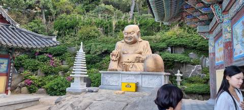 A large laughing Buddha statue sits in a lush Korean temple courtyard framed by traditional tiled roofs and colorful eaves.