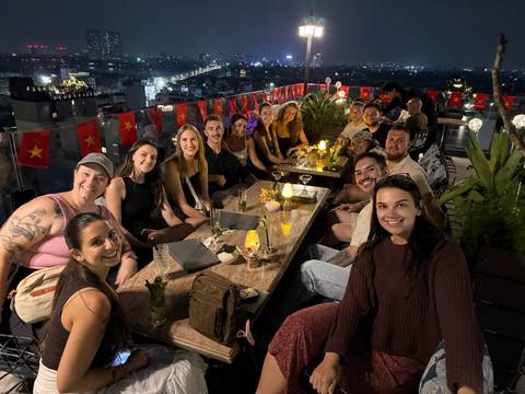 Travellers gather at a rooftop bar at night, city lights and Vietnamese flags in the background.