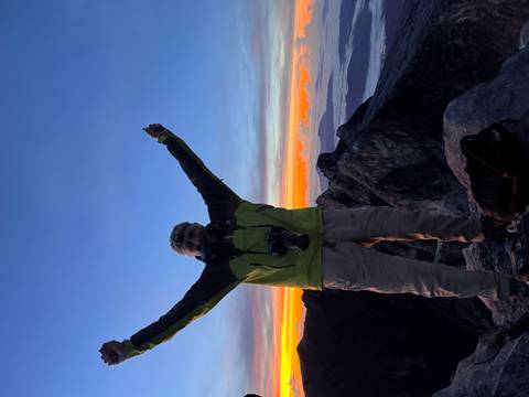 Triumphant climber raising arms on a summit at colourful dawn with valleys blanketed by clouds.