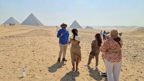 Guide explaining history to a small tour group on the desert plain with Giza pyramids in the distance.