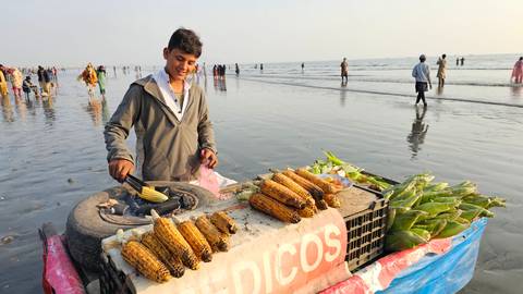 A smiling young vendor roasts corn at his seaside cart while people walk across a wet beach at low tide.