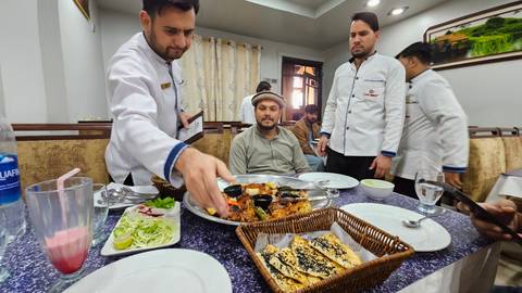 Restaurant staff in white jackets serve a large mixed grill platter to a seated diner inside an eatery.