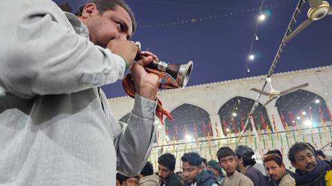 At night a man blows a traditional horn while devotees gather at a festively decorated shrine courtyard.