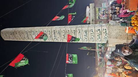 A tall column covered in Urdu script towers over a lively nighttime street market with flags and stalls.