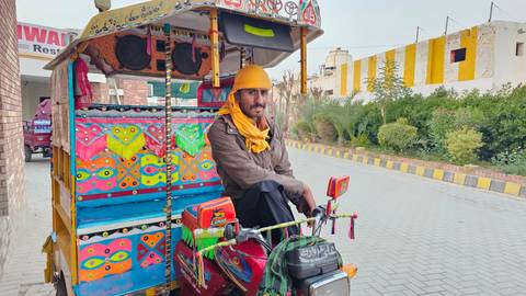 A colorful, artistically painted auto-rickshaw driver poses on his three-wheeler along a paved street.