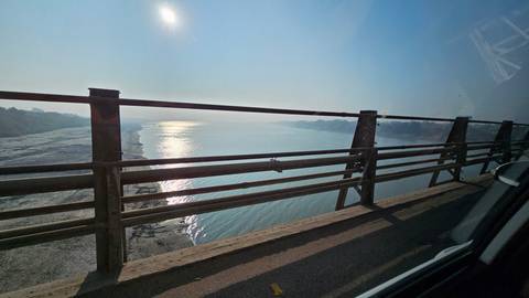 View from a bridge of a wide river reflecting bright sunlight under a hazy blue sky.