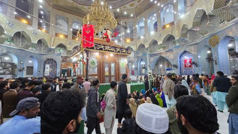 Hundreds of pilgrims crowd a richly tiled shrine interior lit by chandeliers and colorful banners.