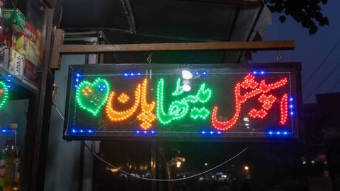 A glowing LED Urdu sign with multicolored lights hangs outside a shop after dusk.