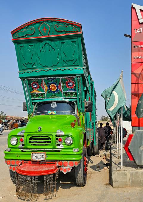 A brightly painted green Pakistani truck decorated with floral motifs stands beside a roadside flag.