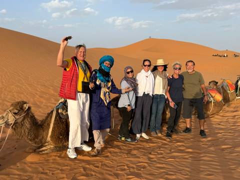 Tour group poses happily beside kneeling camels on golden Sahara dunes in late afternoon light.