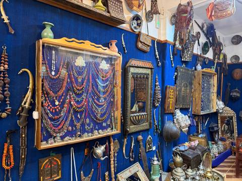 Display cases of colorful Berber jewelry and antique artefacts line a blue-fabric covered wall in a shop.