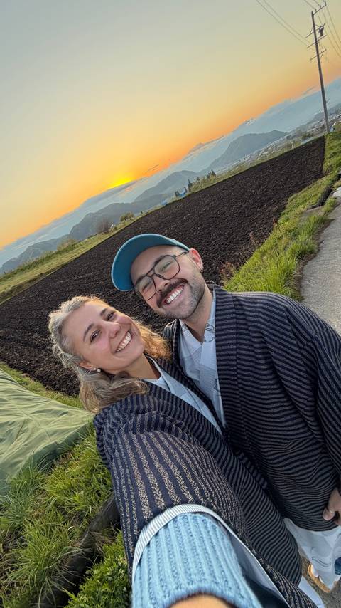 Smiling couple in traditional robes taking selfie with farmland and sunset behind