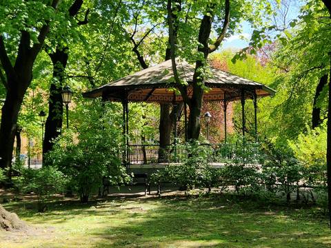 Leafy city park with an ornate iron gazebo shaded by tall trees.