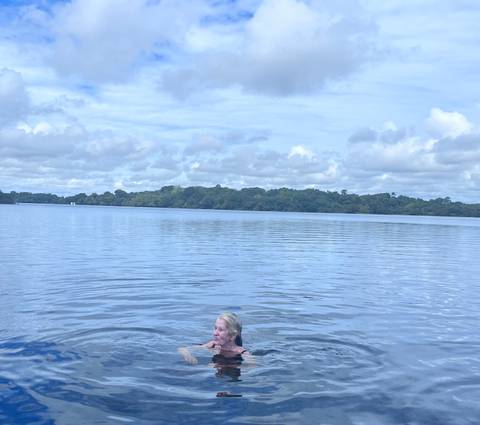 Woman swims in a calm, wide river surrounded by lush rainforest under a cloudy sky.