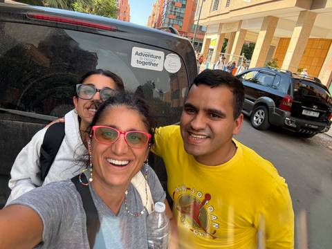 Three smiling travellers take a selfie beside a dusty vehicle on a Moroccan city street.