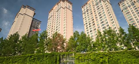 Motion-blurred photo of tall residential high-rises behind a green hedge under blue sky.
