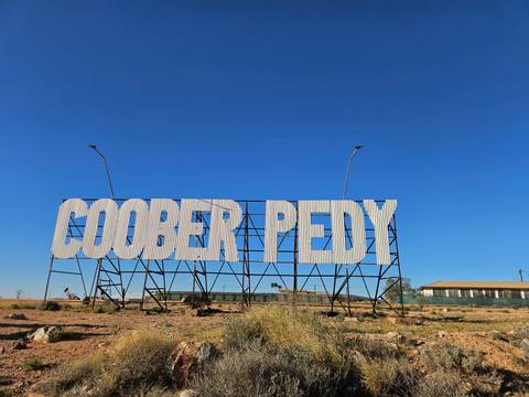 Large white metal sign spelling 'COOBER PEDY' stands against a vivid, cloudless blue sky in the outback.