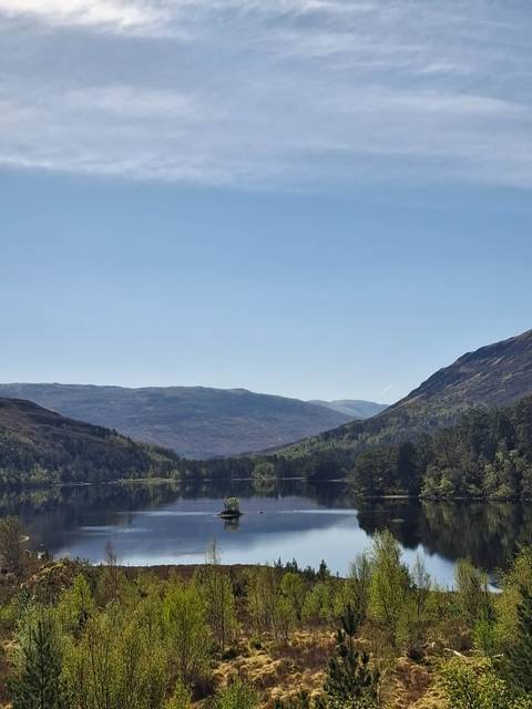 Serene Scottish loch reflecting forested hills beneath a clear blue sky