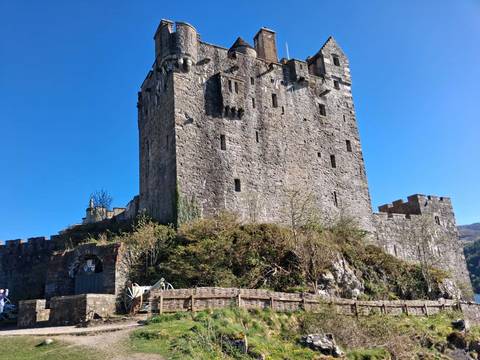 Stone castle rising against a bright blue sky, surrounded by defensive walls and greenery at its base.