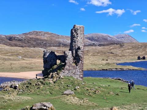 Ruined stone tower on a grassy peninsula beside a blue loch, with rolling brown hills under a clear sky.