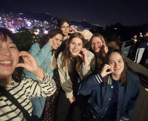 Group of friends smiling and posing for a selfie at night with the illuminated Seoul skyline behind them.