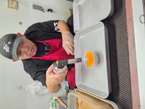 Street-food vendor in apron uses a blowtorch to caramelize a custard dessert.