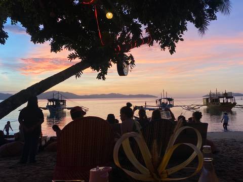 Silhouetted group relaxing under a tree at sunset as colourful boats anchor off an island shore.