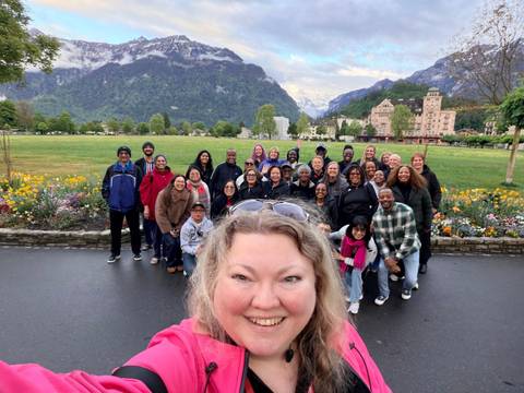 Large tour group poses for a cheerful selfie in a park surrounded by colourful flowers with dramatic Swiss mountains in the background.