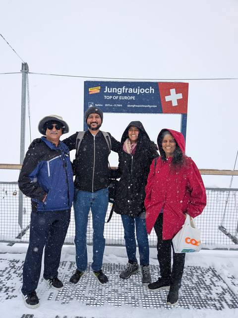 Family bundled in winter jackets smiles beside a 'Top of Europe' sign during falling snow.