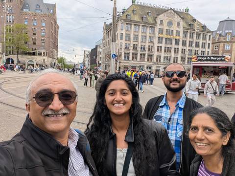 Smiling family takes a selfie in Amsterdam’s bustling Dam Square filled with trams and pedestrians.