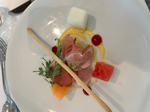 Elegant appetizer of cured ham, fruit cubes, greens and sauce arranged artfully on a white plate.