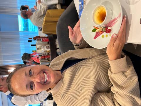 Woman smiling while presenting a small plated dessert at a restaurant table aboard a river cruise.